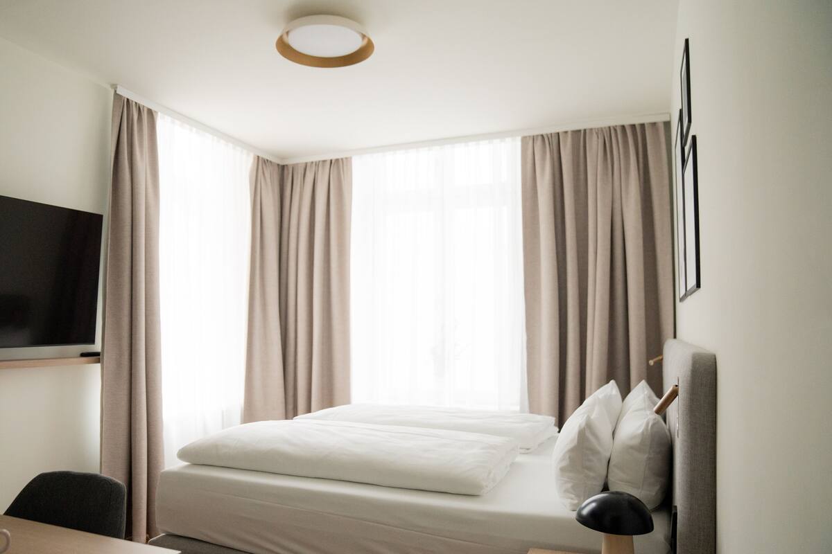 A hotel room with a bed, a desk, a chair, a television, and large windows with curtains.