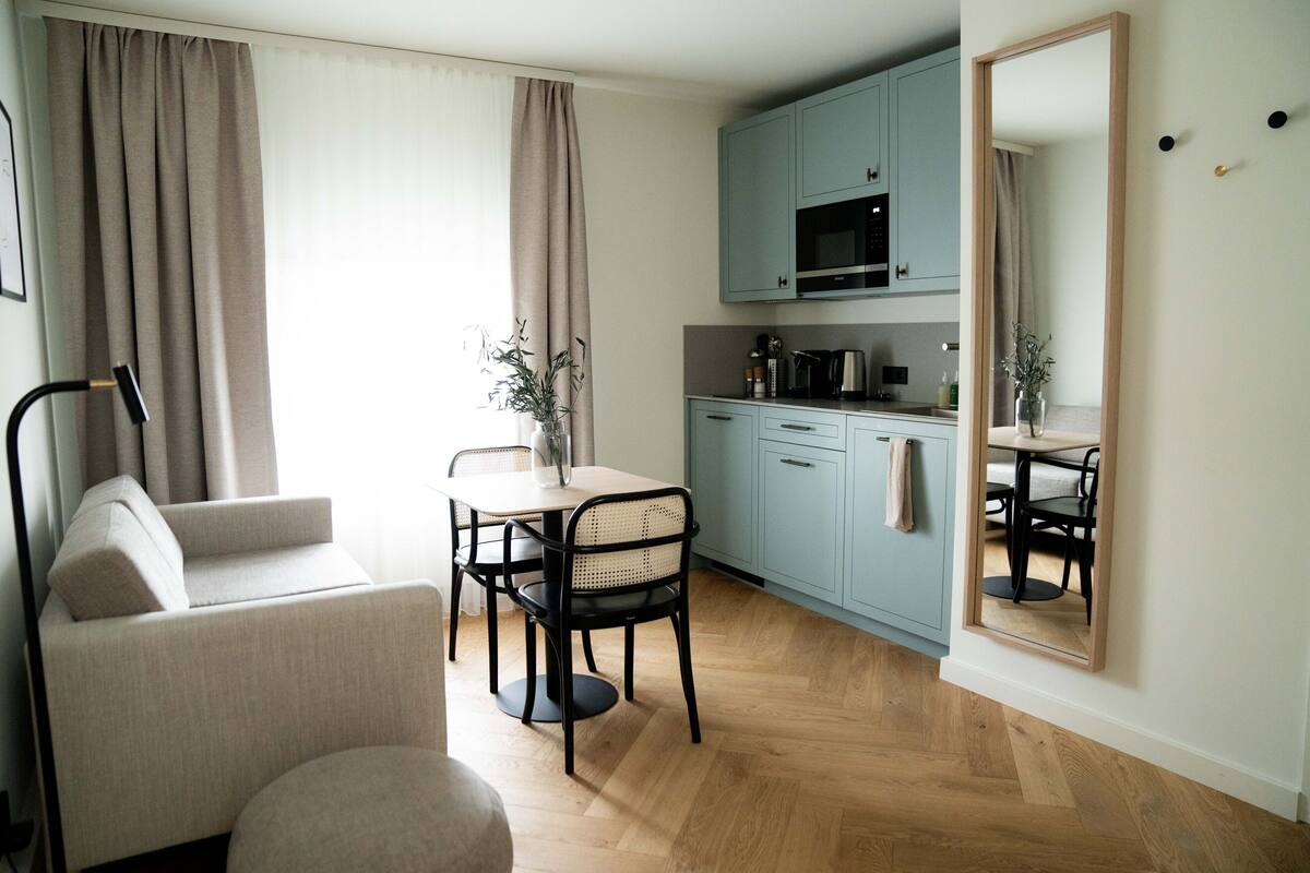 A compact living space with a sofa, dining table, chairs, a kitchen area with cabinets and appliances, and a large mirror.