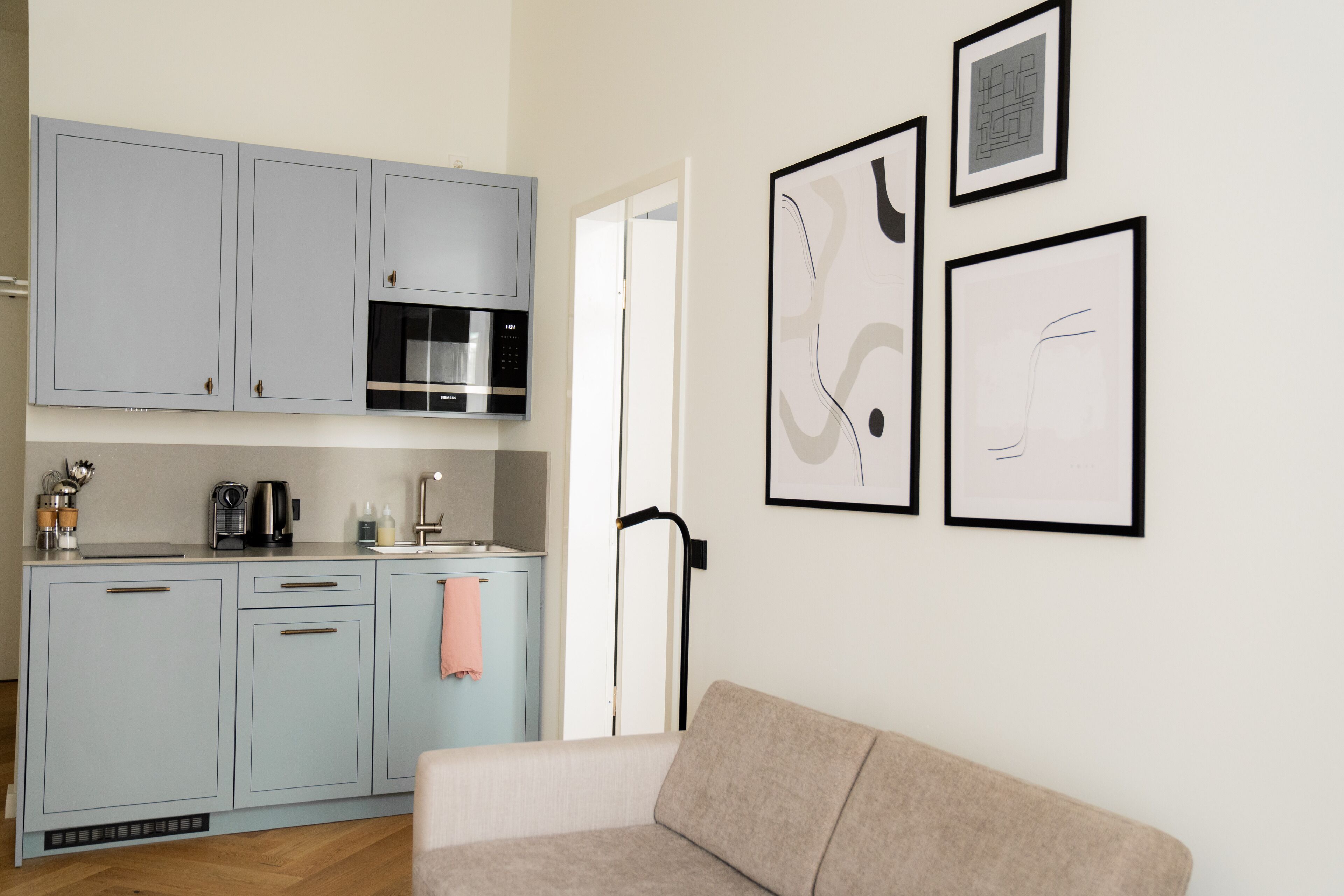 A modern kitchen with grey cabinets, a microwave, and a coffee maker. There is a beige sofa in the foreground and abstract art on the wall.