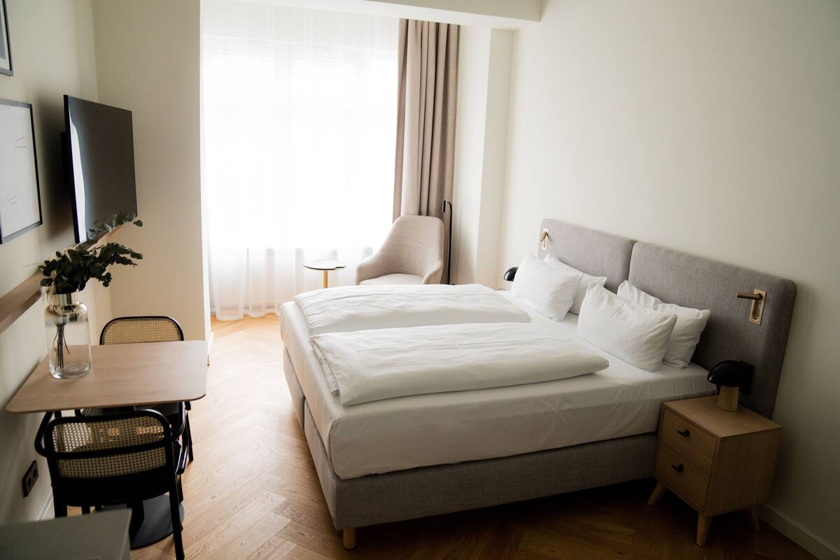 A hotel room with a bed, bedside table, chair, desk, and a TV on the wall.