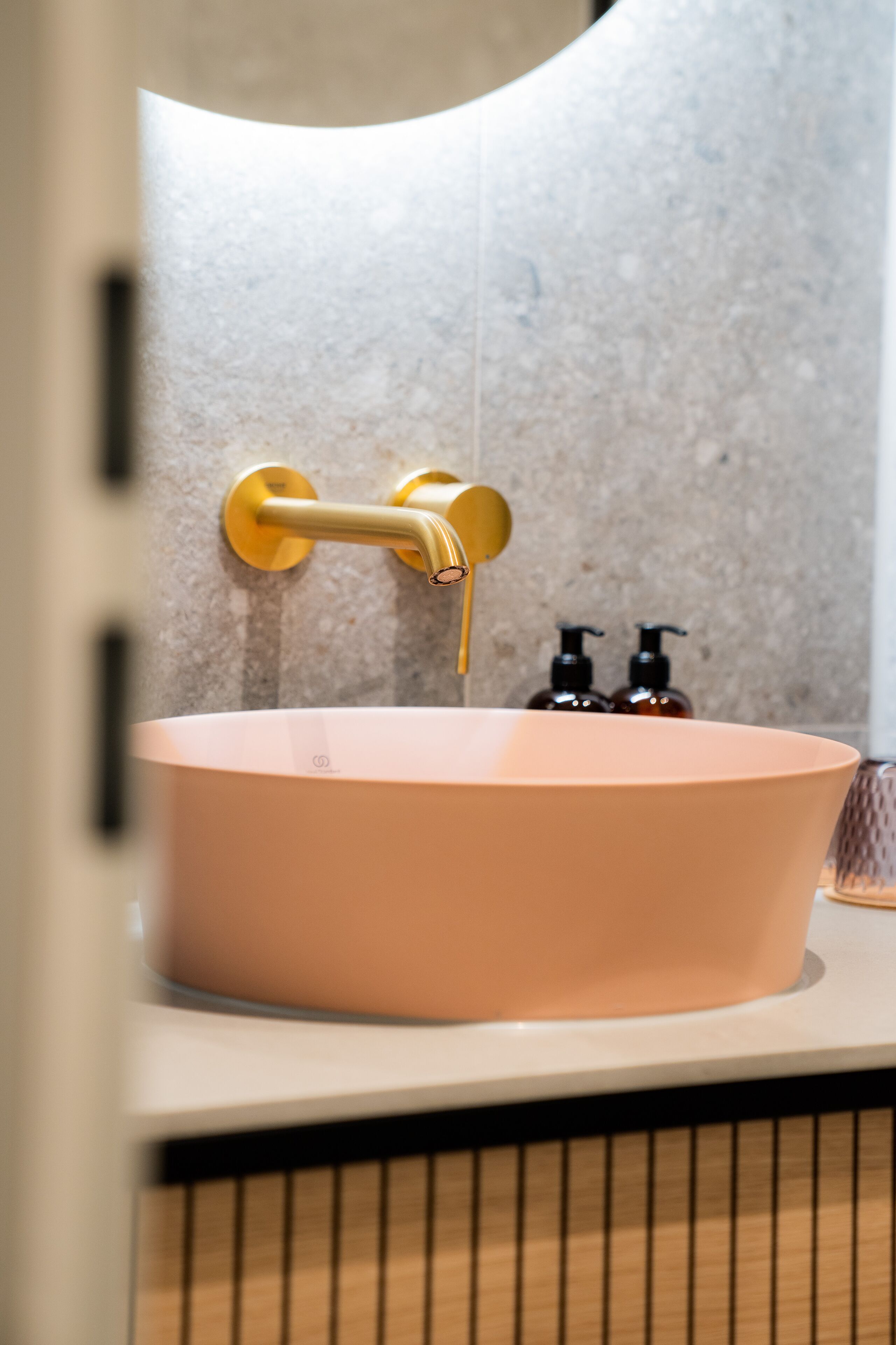 A bathroom with a pink sink, gold faucet, and two soap dispensers on the wall.