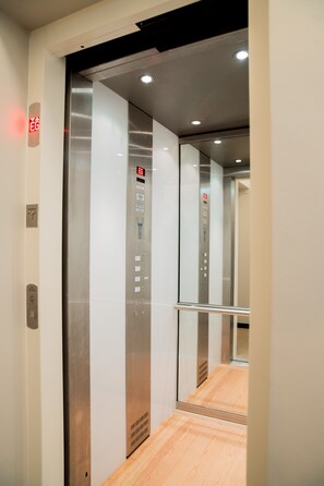 Interior de elevador com paredes metálicas, piso de madeira e painel de controle com botões de emergência.