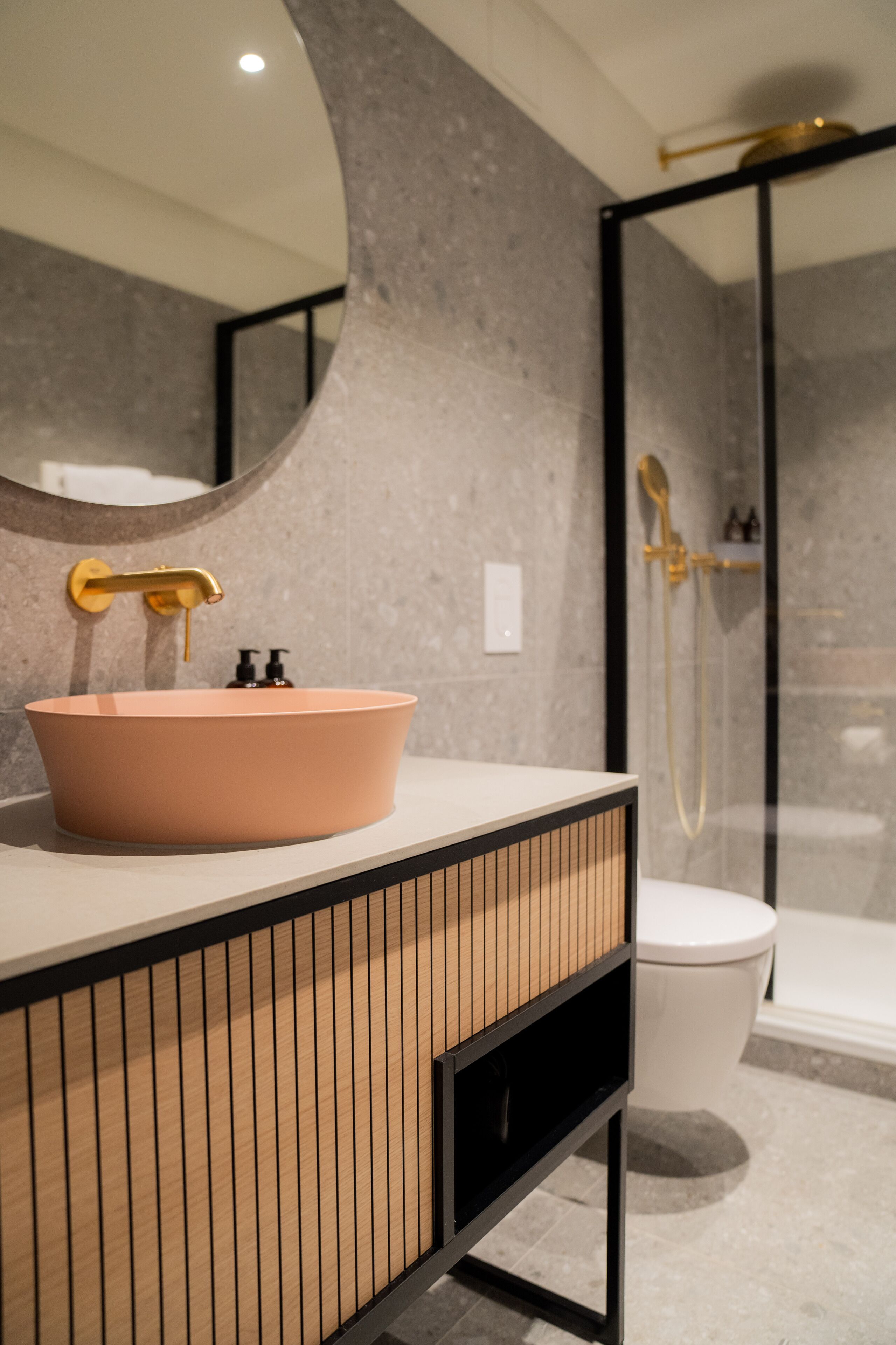 Une salle de bain avec un lavabo rond de couleur rose, un meuble en bois à panneaux, des toilettes blanches et une douche séparée par une paroi en verre.