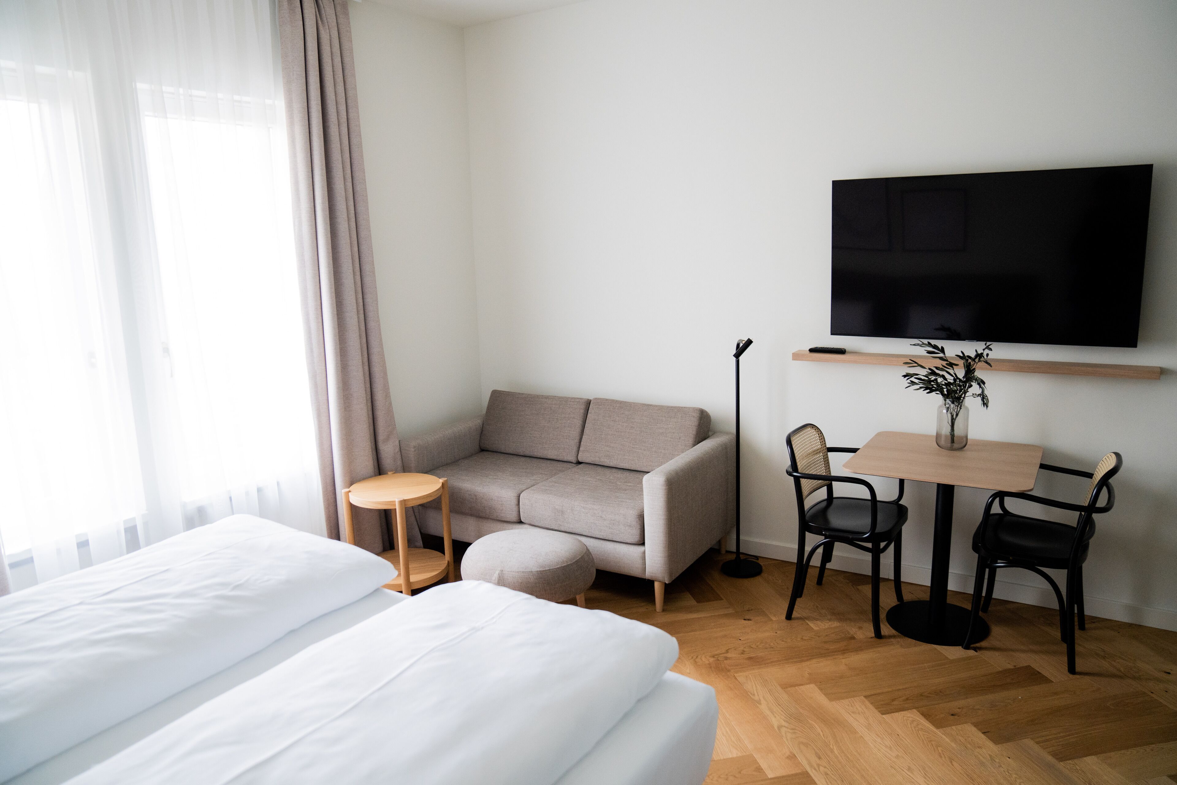 A hotel room with a bed, a sofa, a small table with chairs, a TV mounted on the wall, and a window with curtains.