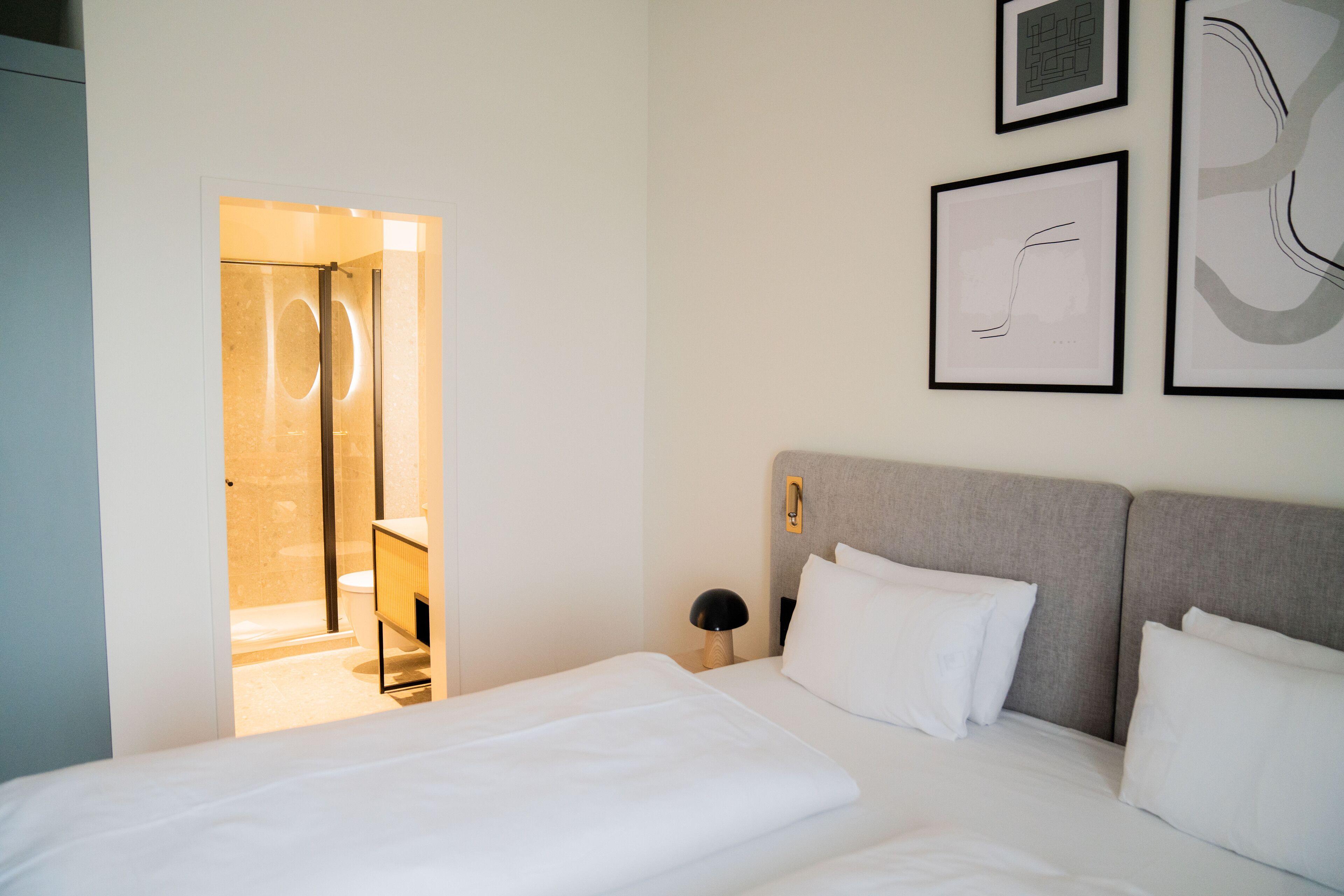 Une chambre d’hôtel moderne, équipée d’un lit, d’une table de chevet et d’une lampe fixée au mur. On peut apercevoir la salle de bain à travers une porte ouverte.