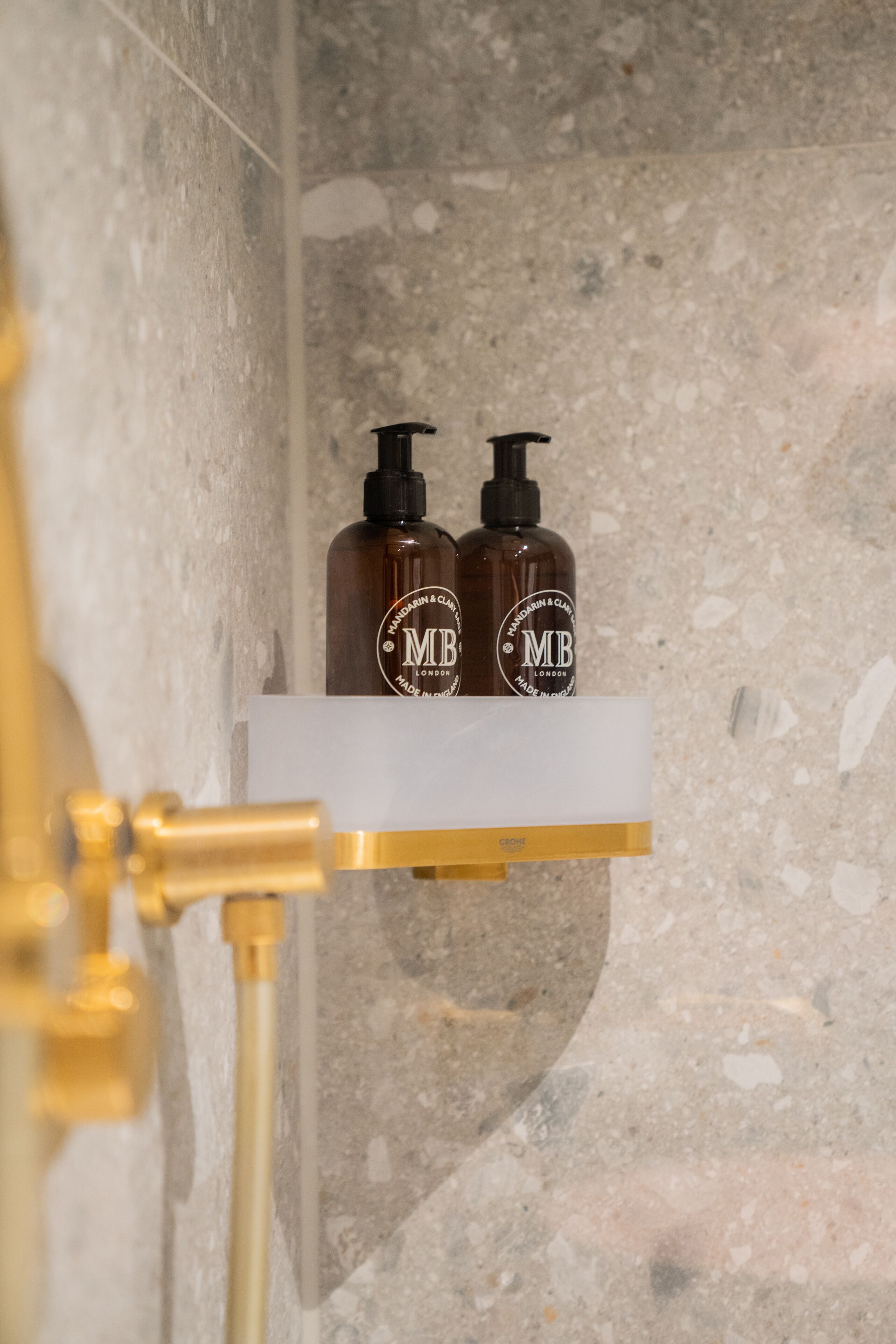 A bathroom with a marble wall, a white shelf holding two brown bottles labeled 'MB London', and a gold faucet.
