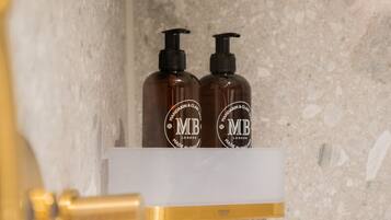 A bathroom with a marble wall, a white shelf holding two brown bottles labeled 'MB London', and a gold faucet.
