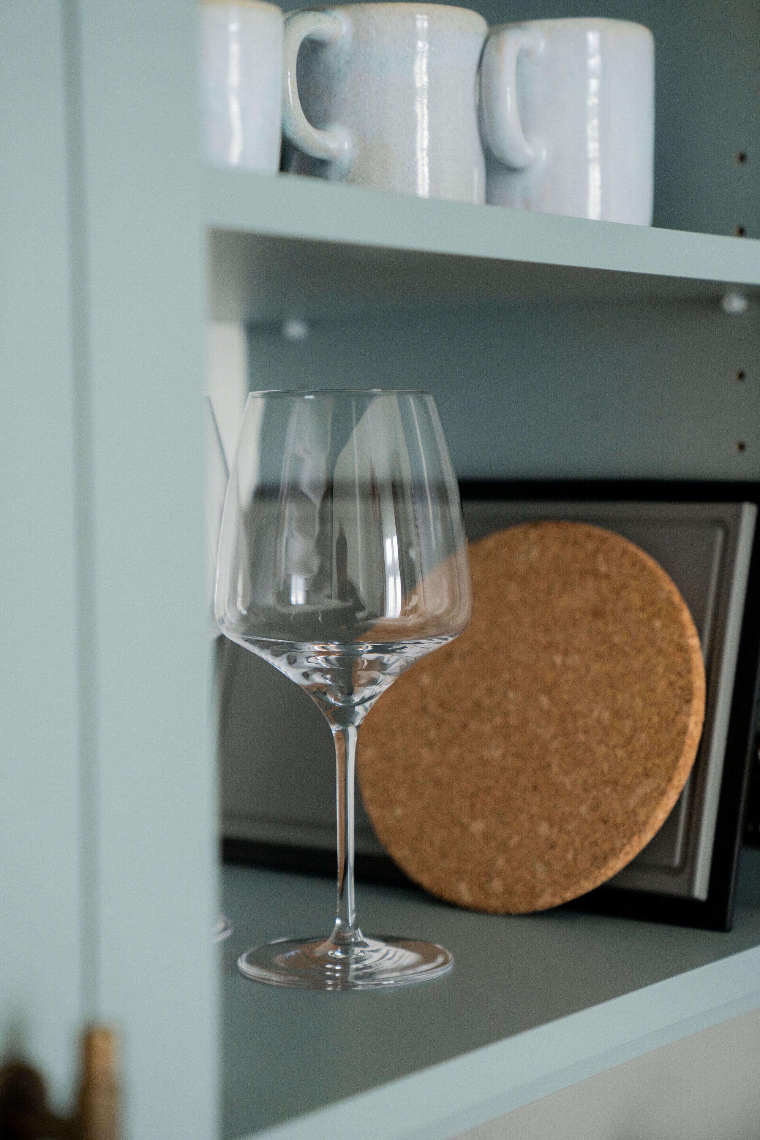 A clear glass wine glass on a shelf with a cork coaster and white ceramic mugs in the background.