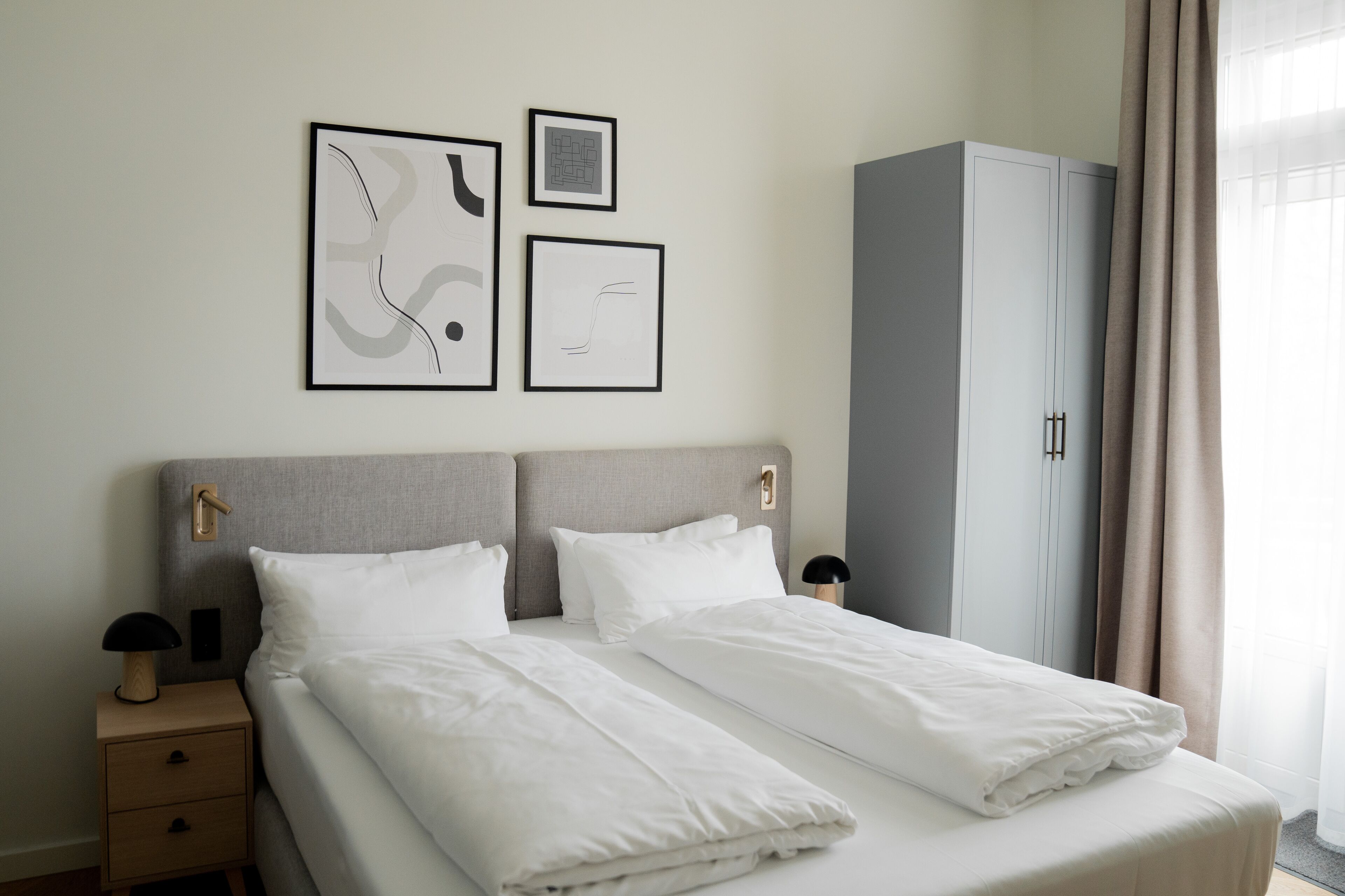 Una cama bien hecha con sábanas blancas, un cabecero gris y dos mesitas de noche con lámparas. Se observan obras de arte enmarcadas en la pared y un armario a la derecha.