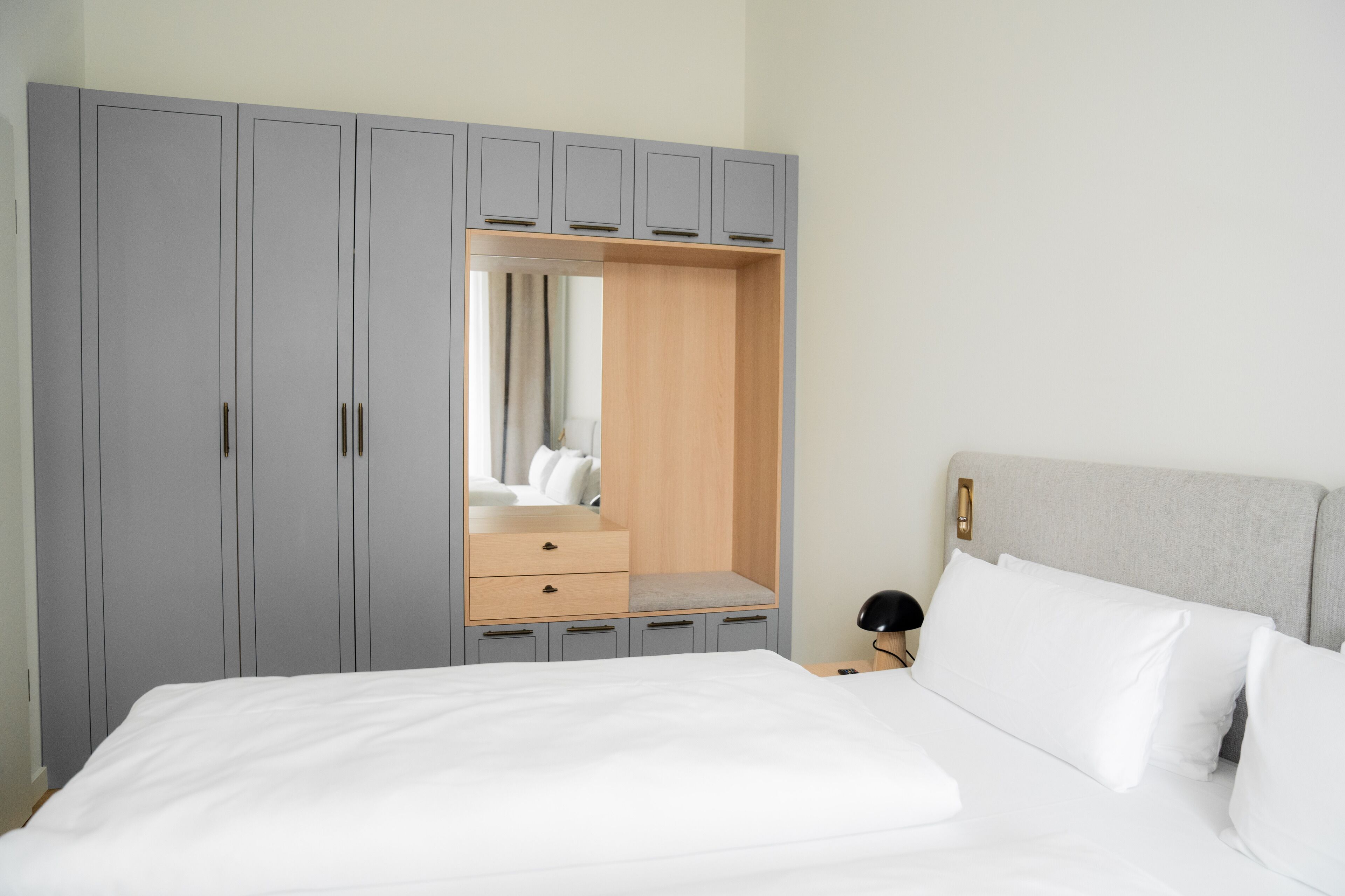 A bedroom with a bed, a grey wardrobe with a mirror, and a bedside table with a lamp.