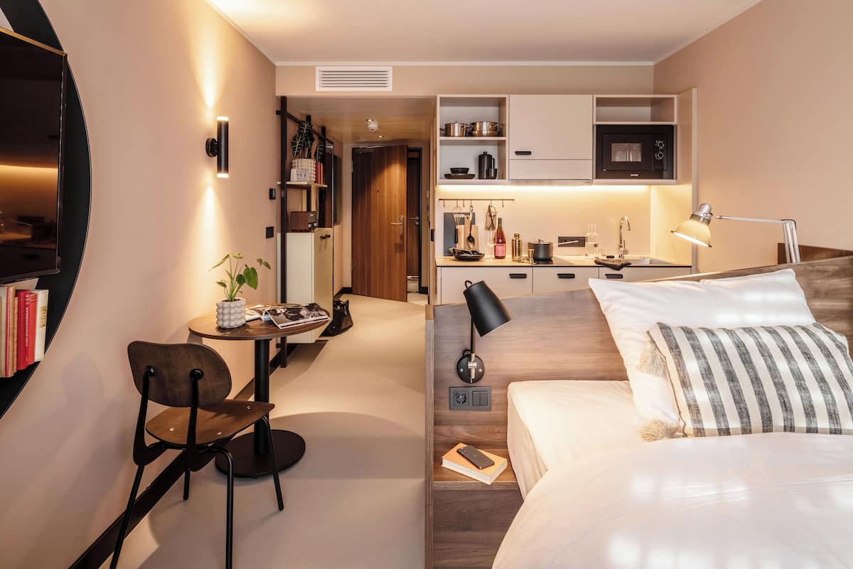 Una habitación de hotel compacta con cama, mesita de noche, silla, kitchenette y un pequeño comedor.