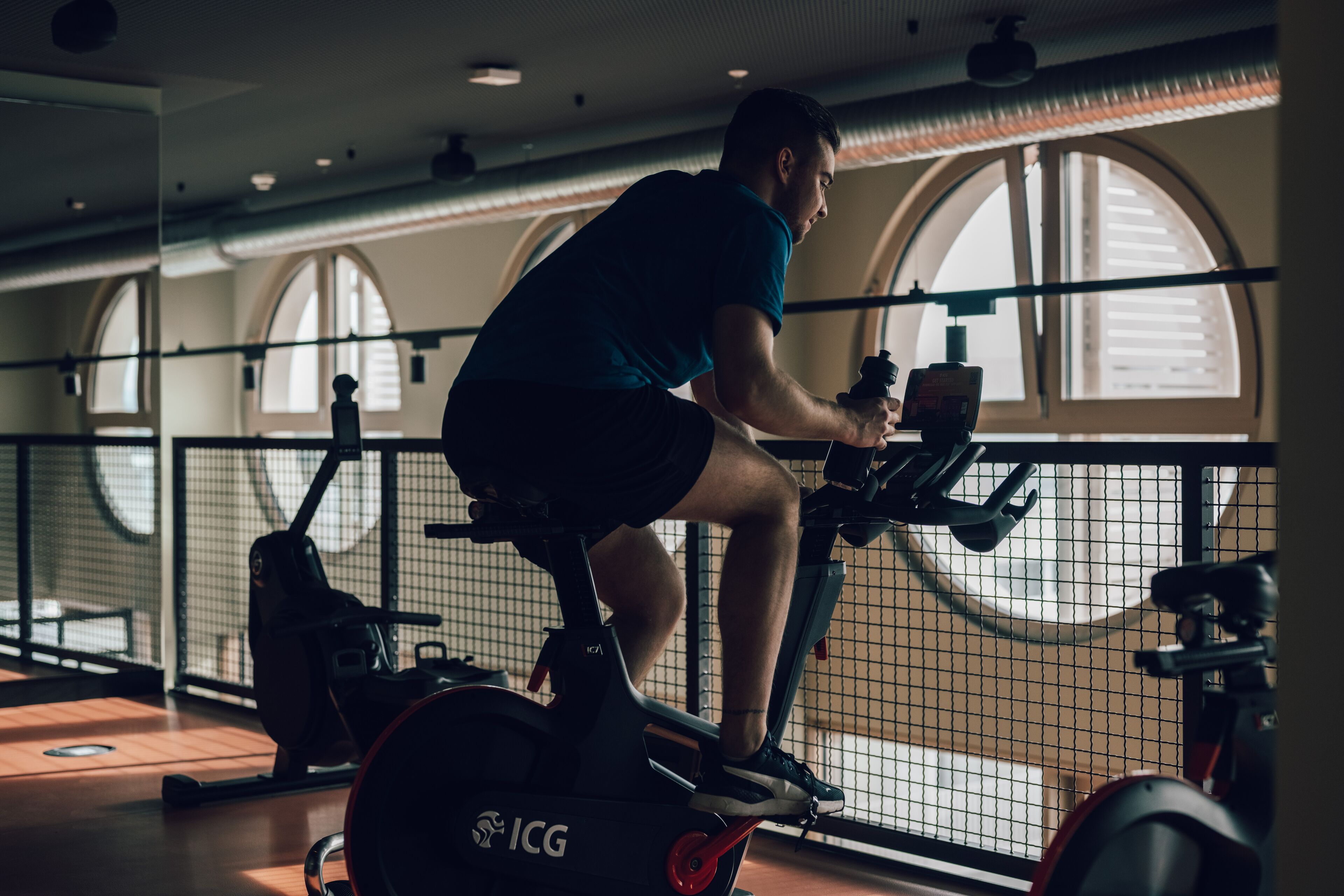 A person cycling on an ICG stationary bike in a gym with large windows and a wooden floor.
