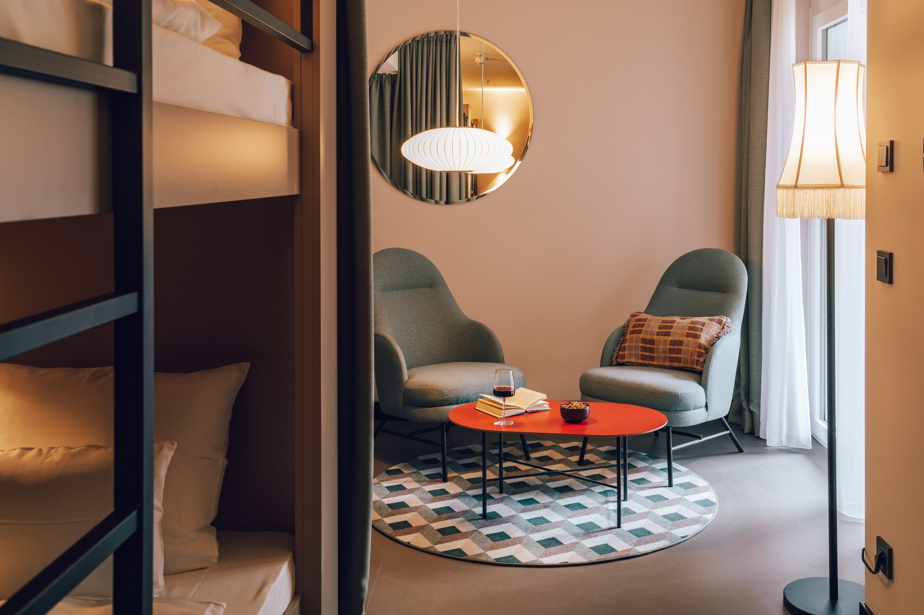 A hotel room with a bunk bed, two armchairs, a small round table with a book and glass, a patterned rug, a round mirror, and a floor lamp.