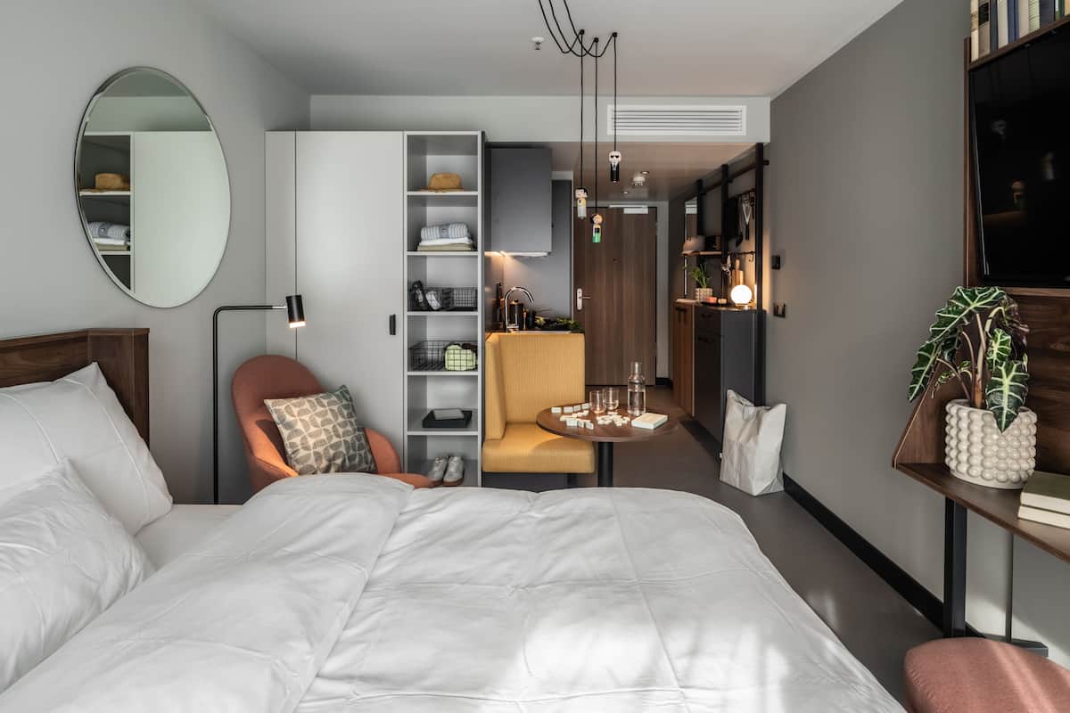 Una habitación de hotel moderna con una cama grande, una zona de estar con una silla, una mesita, un armario y un espejo.