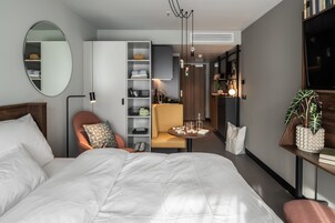 Ein modernes Hotelzimmer mit einem großen Bett, einer Sitzecke mit einem Sessel, einem kleinen Tisch, einem Kleiderschrank und einem Spiegel.