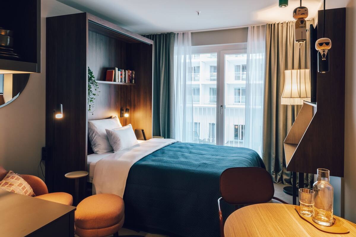 Habitación de hotel con una cama grande, un escritorio, una silla, una mesita, una estantería y vistas a la ciudad a través de la ventana.