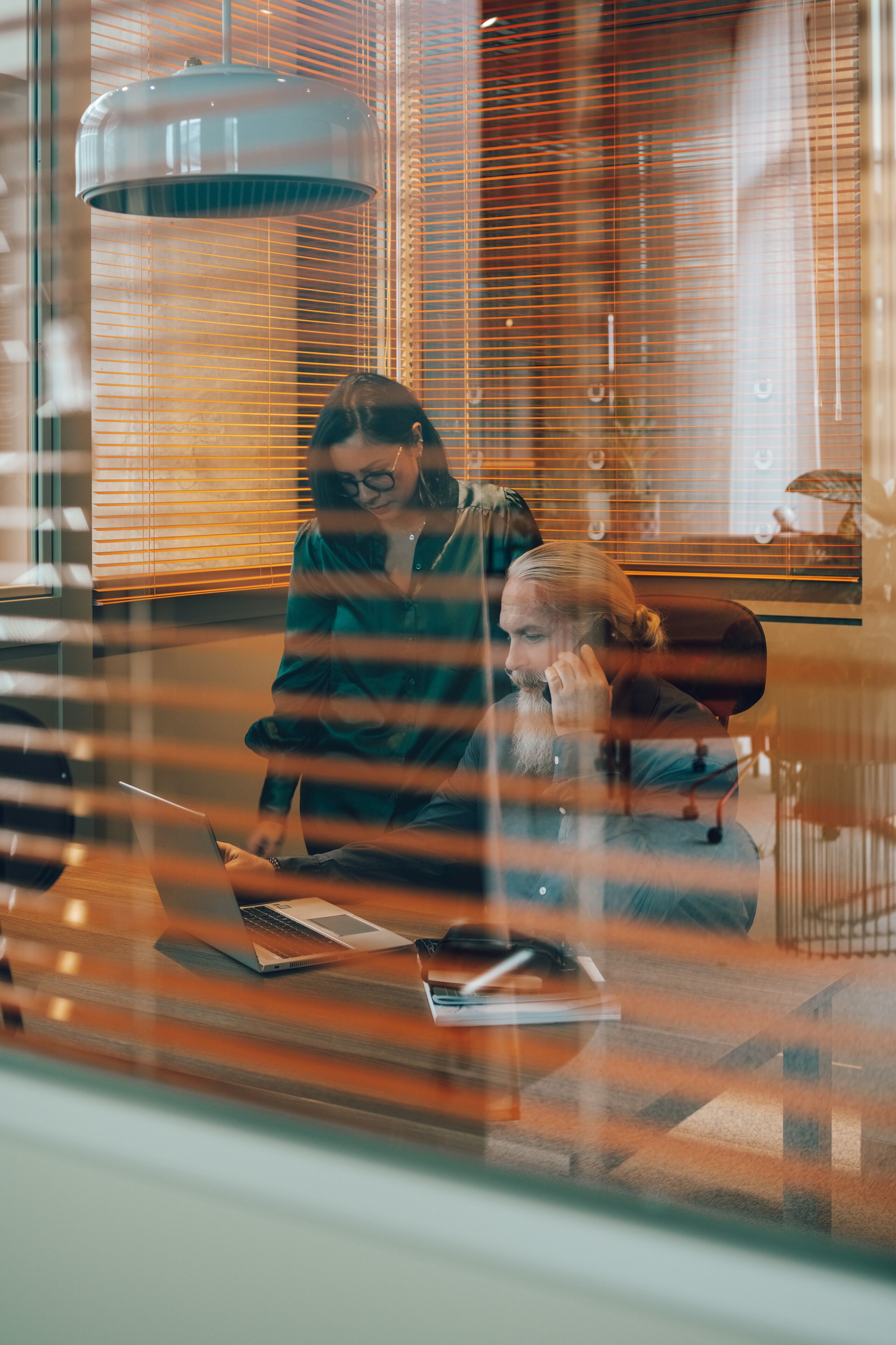 Una persona sentada a una mesa con una computadora portátil, otra persona sentada al fondo y un ventanal con persianas.