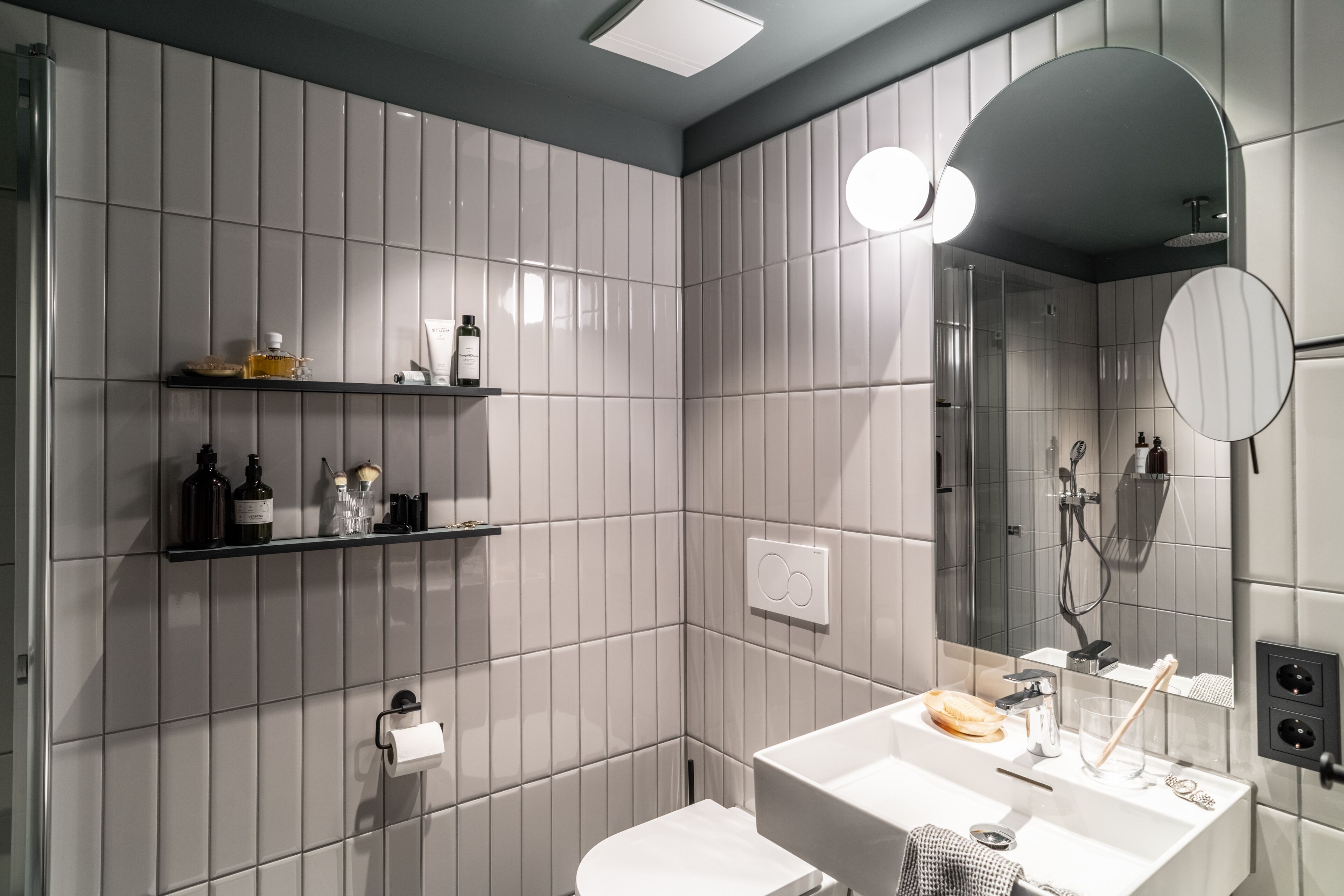 Ein modernes Badezimmer mit Toilette, Waschbecken mit Spiegel und Duschbereich. Es gibt Regale mit Pflegeprodukten und einen Handtuchhalter.