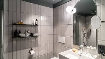 Ein modernes Badezimmer mit Toilette, Waschbecken mit Spiegel und Duschbereich. Es gibt Regale mit Pflegeprodukten und einen Handtuchhalter.
