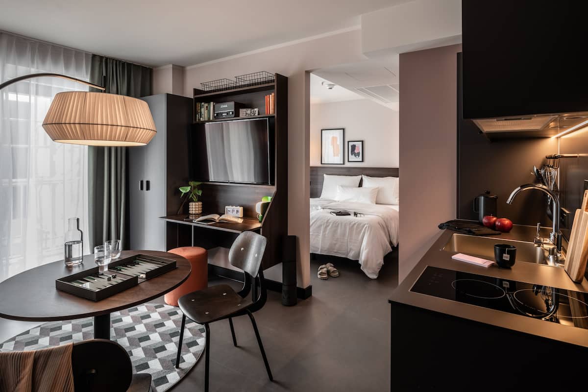 Una habitación de hotel moderna con comedor, cama, kitchenette y un amplio ventanal con cortinas.