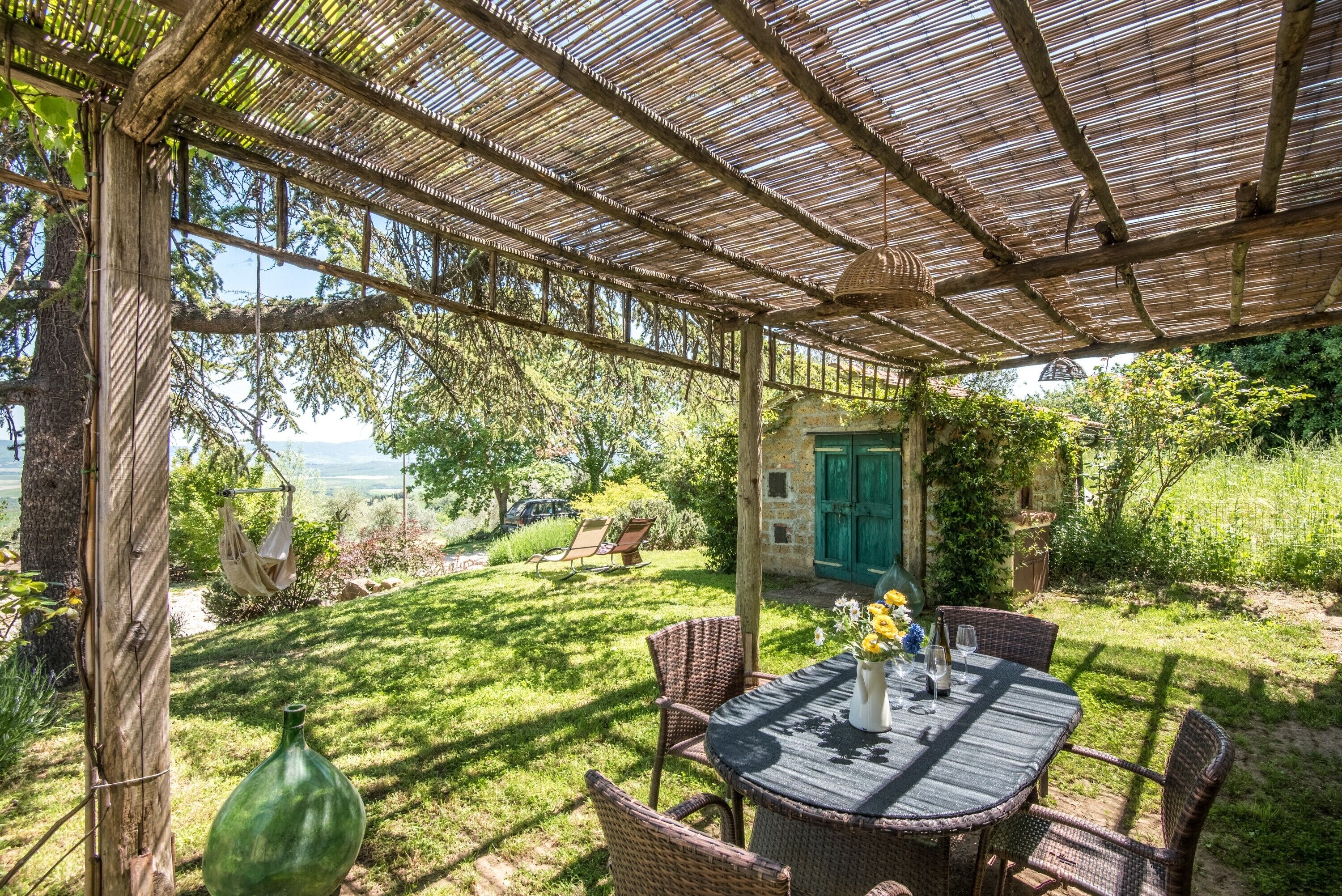 Un espace extérieur aménagé, abrité et doté de meubles en osier et d’une table dressée pour le repas, entouré d’une végétation luxuriante et d’un bâtiment en pierre avec une porte verte.