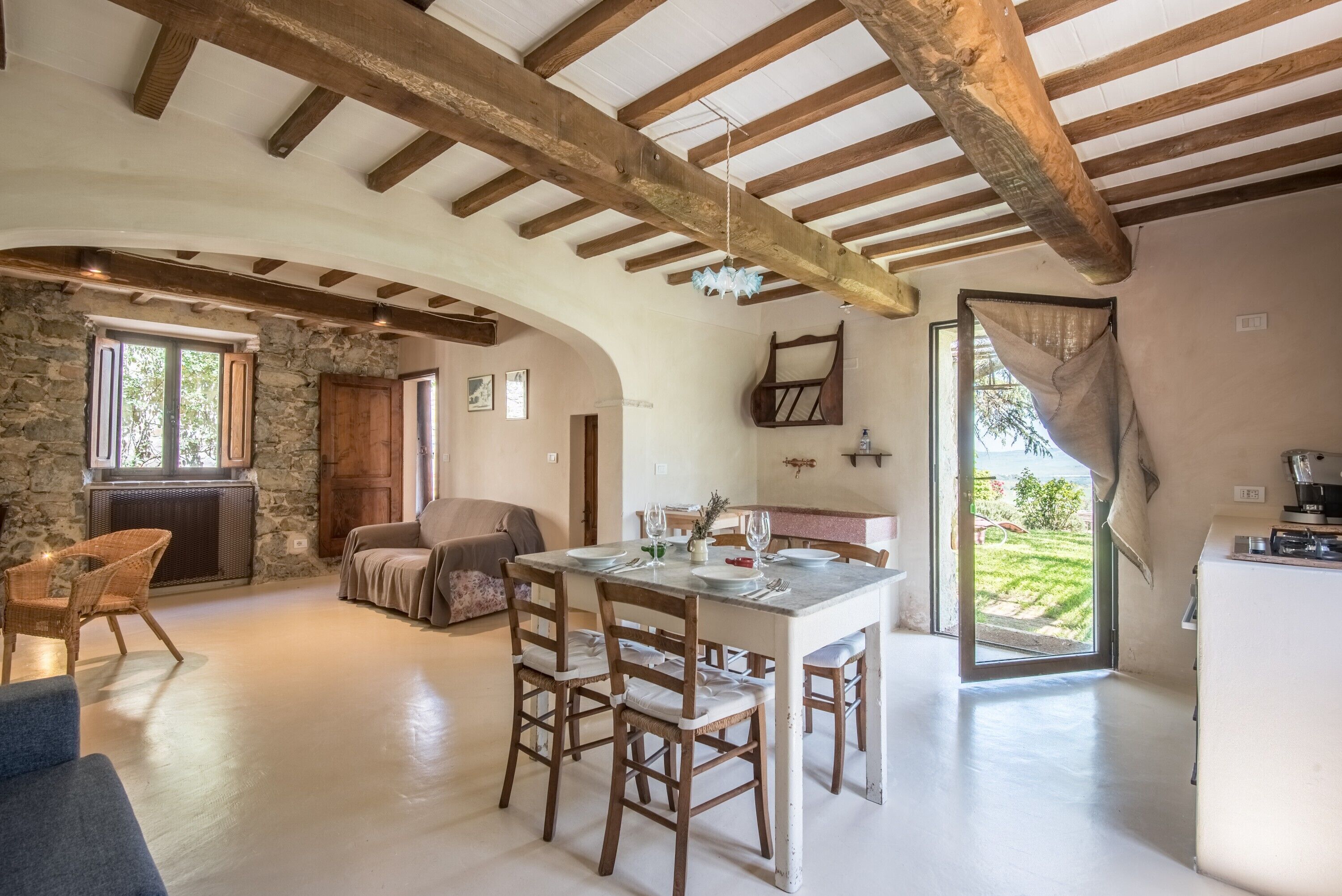 Une pièce spacieuse avec un mur en pierre, des poutres apparentes, une table à manger avec des chaises, un canapé et une vue sur un jardin.