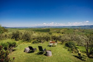 A grassy outdoor area with two chairs and a table, surrounded by trees and hills in the background.