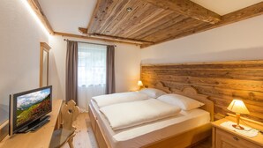 Quarto com teto de madeira, cama com roupa de cama branca, TV sobre uma escrivaninha de madeira e mesa de cabeceira com abajur.