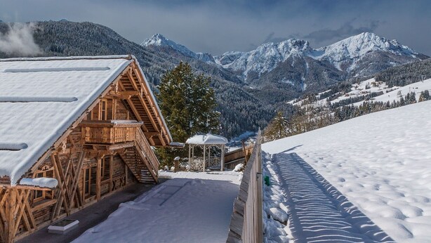 Een houten chalet met een met sneeuw bedekte dak, een houten terras en uitzicht op besneeuwde bergen.