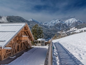 A wooden chalet with a snow-covered roof, a wooden deck, and a view of snow-covered mountains.