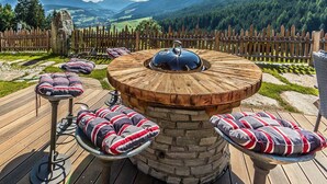 Een houten zitgedeelte buiten, met een stenen tafel, comfortabele stoelen en uitzicht op bergen.