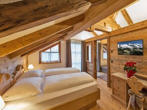Une chambre mansardée chaleureuse avec un plafond en bois, un lit recouvert de draps blancs, une commode et un tableau représentant un paysage de montagne.