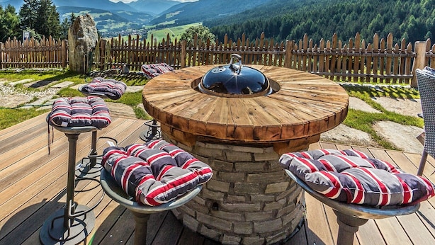 A wooden outdoor seating area with a stone table, cushioned chairs, and a view of mountains. - Apartment Piz da Peres - Geiselsberg with Mountain View, Pool, Sauna, Wi-Fi & Garden (Olang)