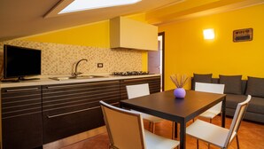 Eine moderne Küche mit integriertem Fernseher, ein Essbereich mit Tisch und Stühlen und ein Sofa in einem gelb gestrichenen Wohnraum.
