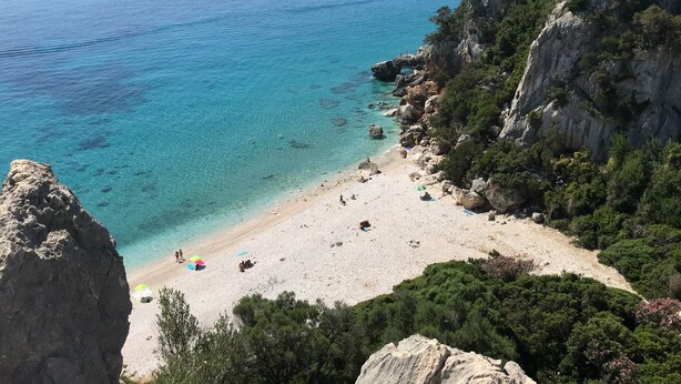 En kustnära strand med klart, turkosfärgat vatten, klippiga branter och gles vegetation.