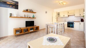 A modern living room with a TV, wooden shelves, a dining table with chairs, and a kitchen area with white cabinets and a tiled backsplash. - Holiday Apartment "Casa Antonella" with Garden, Balcony & Air Conditioning (Orosei)