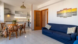 A modern living room with a blue sofa, wooden dining table, and chairs. A kitchen is visible in the background with white cabinets and a stove. - Apartment "Casa Riviera" with Balconies and Wi-Fi (Donnalucata)