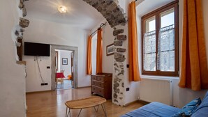 A room with a stone wall, wooden floor, a blue sofa, a wicker chair, a wooden dresser, and a window with orange curtains.