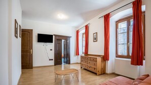 Une pièce avec un plancher en bois, un canapé, une commode en bois, un téléviseur fixé au mur et une fenêtre avec des rideaux rouges.