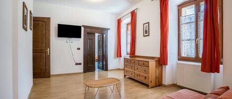 A room with a wooden floor, a sofa, a wooden dresser, a television mounted on the wall, and a window with red curtains.