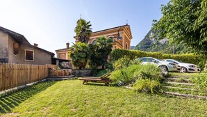 Un jardin avec une terrasse en bois, une pelouse, une voiture et un bâtiment dont l’arrière-plan est constitué d’une chaîne de montagnes.