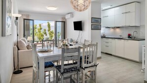 A modern kitchen with white cabinets, a dining area with a table and chairs, a TV mounted on the wall, and a large window with a view of greenery. - Holiday Home "Alegria" close to the Beach with Garden & Wi-Fi (Puerto del Carmen)