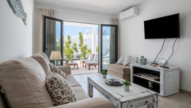 A living room with a sofa, coffee table, TV, and a sliding glass door leading to an outdoor area with plants. - Holiday Home "Alegria" close to the Beach with Garden & Wi-Fi (Puerto del Carmen)