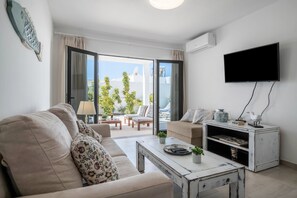 A living room with a sofa, coffee table, TV, and a sliding glass door leading to an outdoor area with plants.