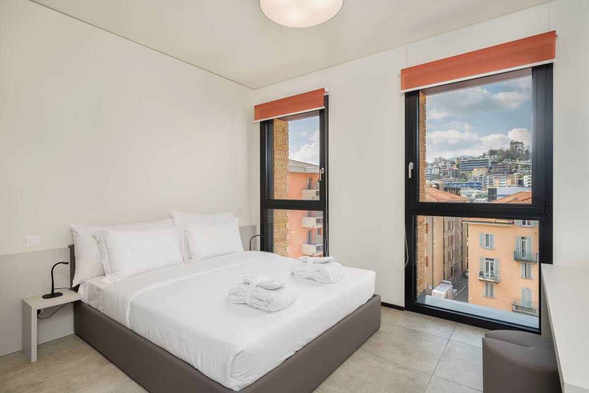 Ein modernes Hotelzimmer mit einem großen Bett, zwei Fenstern mit Jalousien, einem Nachttisch und Blick auf die Stadt.
