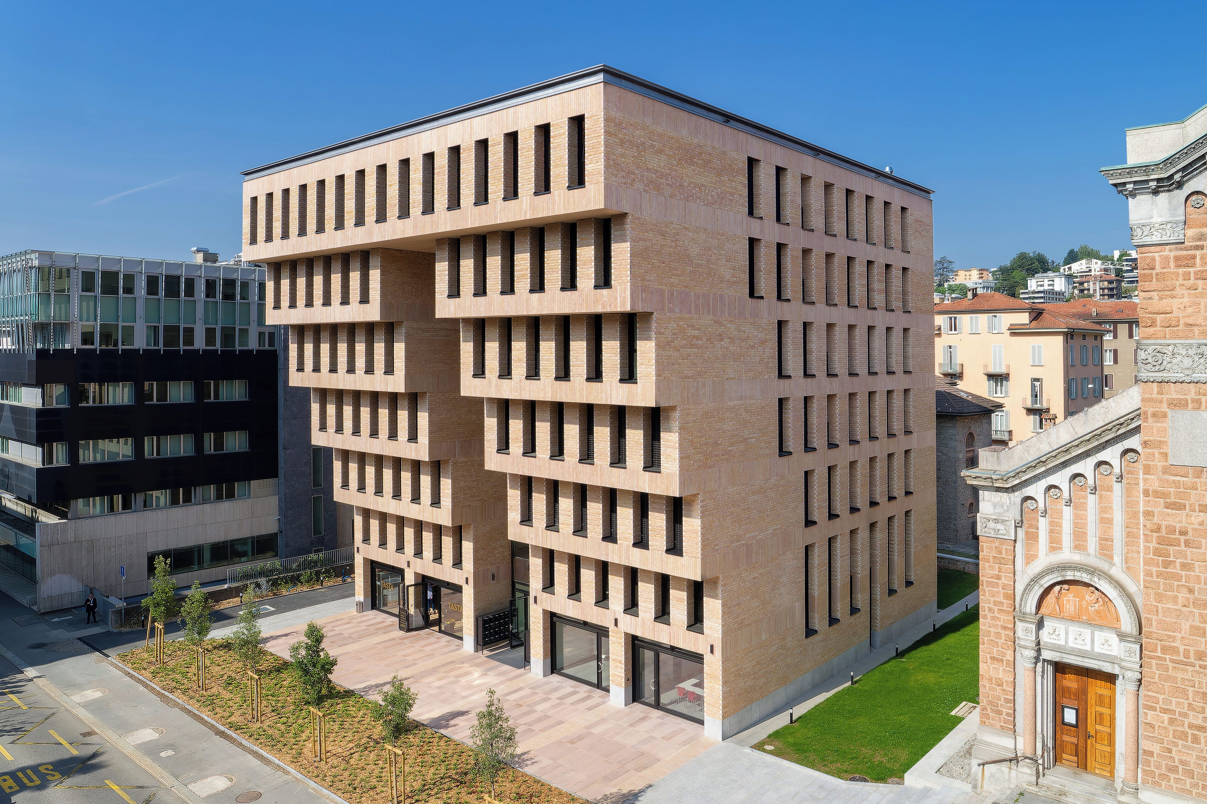 Un immeuble à plusieurs étages doté d’une architecture moderne, caractérisé par un mélange de façades en verre et en briques, et un patio central agrémenté d’arbres.