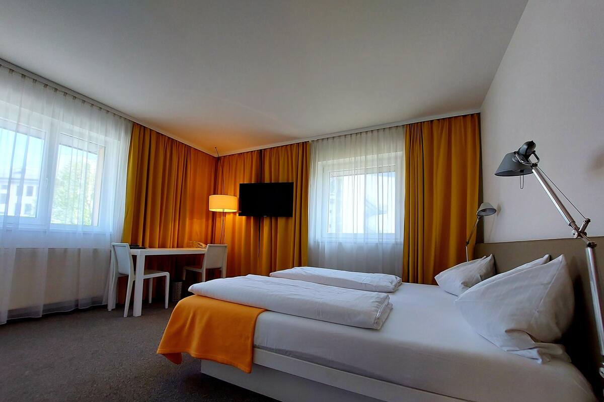 A hotel room with two beds, a desk, a chair, a TV, and a lamp.