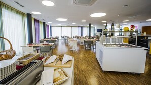 A spacious dining area with multiple tables and chairs, a buffet station, and a kitchen area. - STAY.inn Comfort Art Hotel Schwaz (Schwaz)