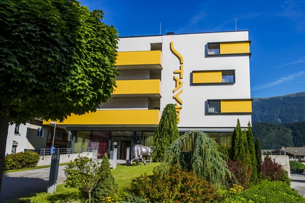A modern building with a yellow and white facade, a large tree in the foreground, and a mountain in the background.