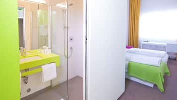 A hotel room with a glass-enclosed shower, a bright yellow sink, a white bed with green bedding, and a window with yellow curtains.